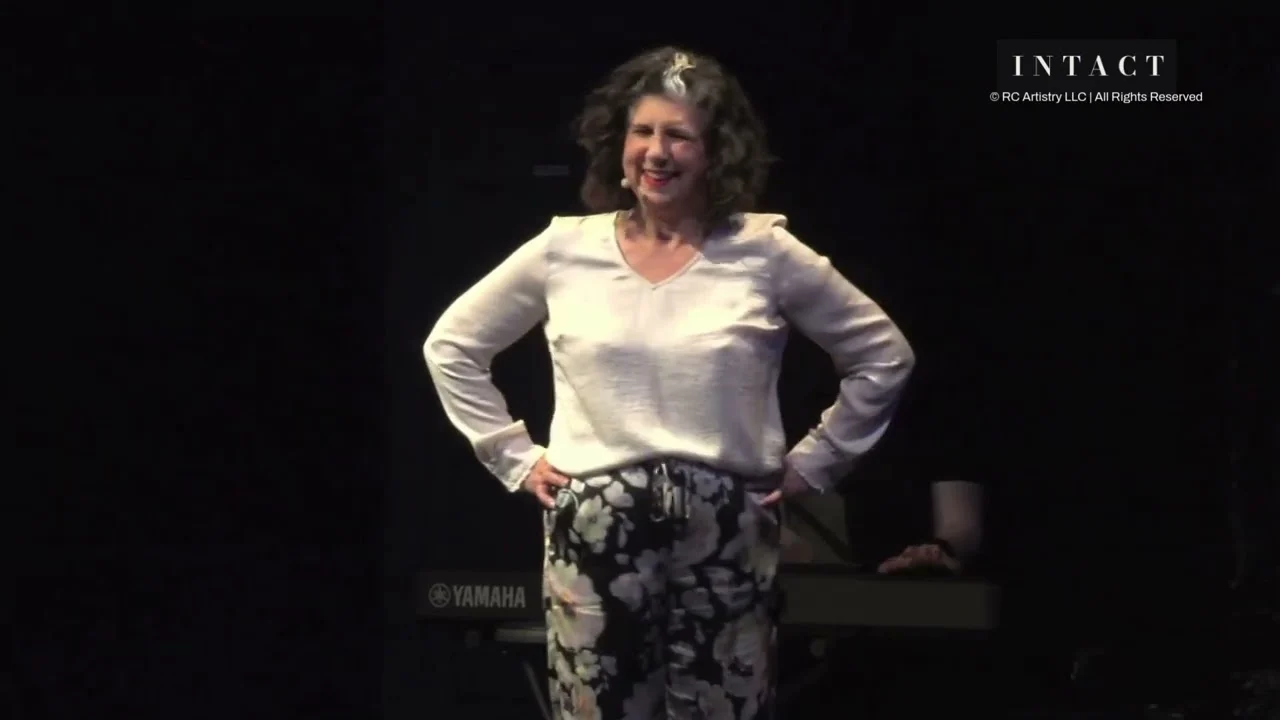 Robin Colucci in a white blouse, hands on her hips, smiling mid-song on the Theatre Row stage
